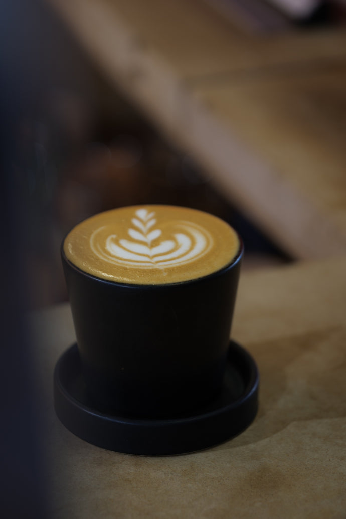 A black ceramic Ritual Coffee branded beaker with saucer on a table, containing a cup of coffee with latte art on top.