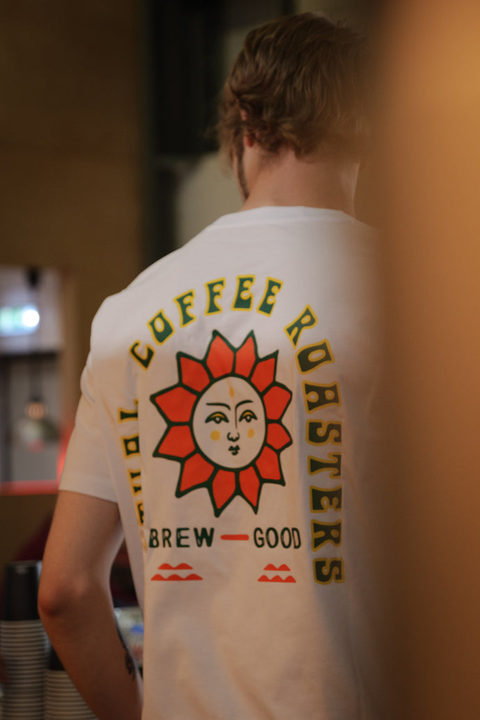 A person wearing a Ritual Coffee branded white crew neck t-shirt with a green and red sun design and the text 'COFFEE ROASTERS BREW GOOD'.