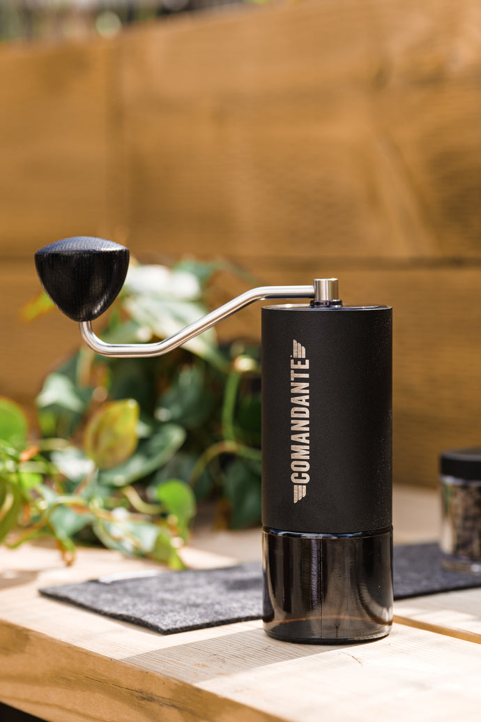 A black Comandante C40 Nitro Blade Grinder with a silver crank, placed on a table with a plant in the background.